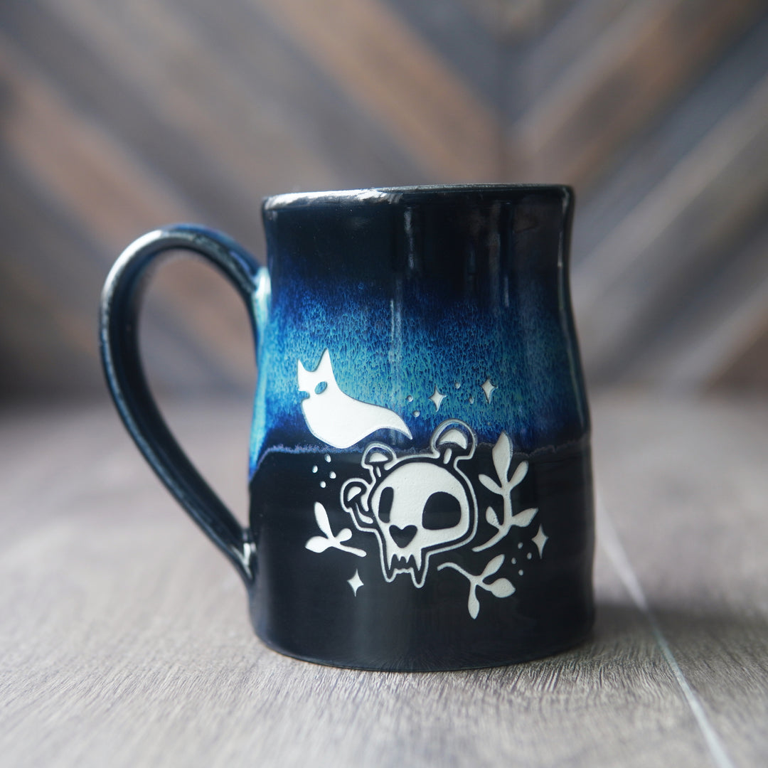 A dark blue and black ceramic mug with a glossy finish, decorated with white illustrations of a cat ghost, a skull, and small stars, set on a wooden surface with a blurred wood-paneled background.