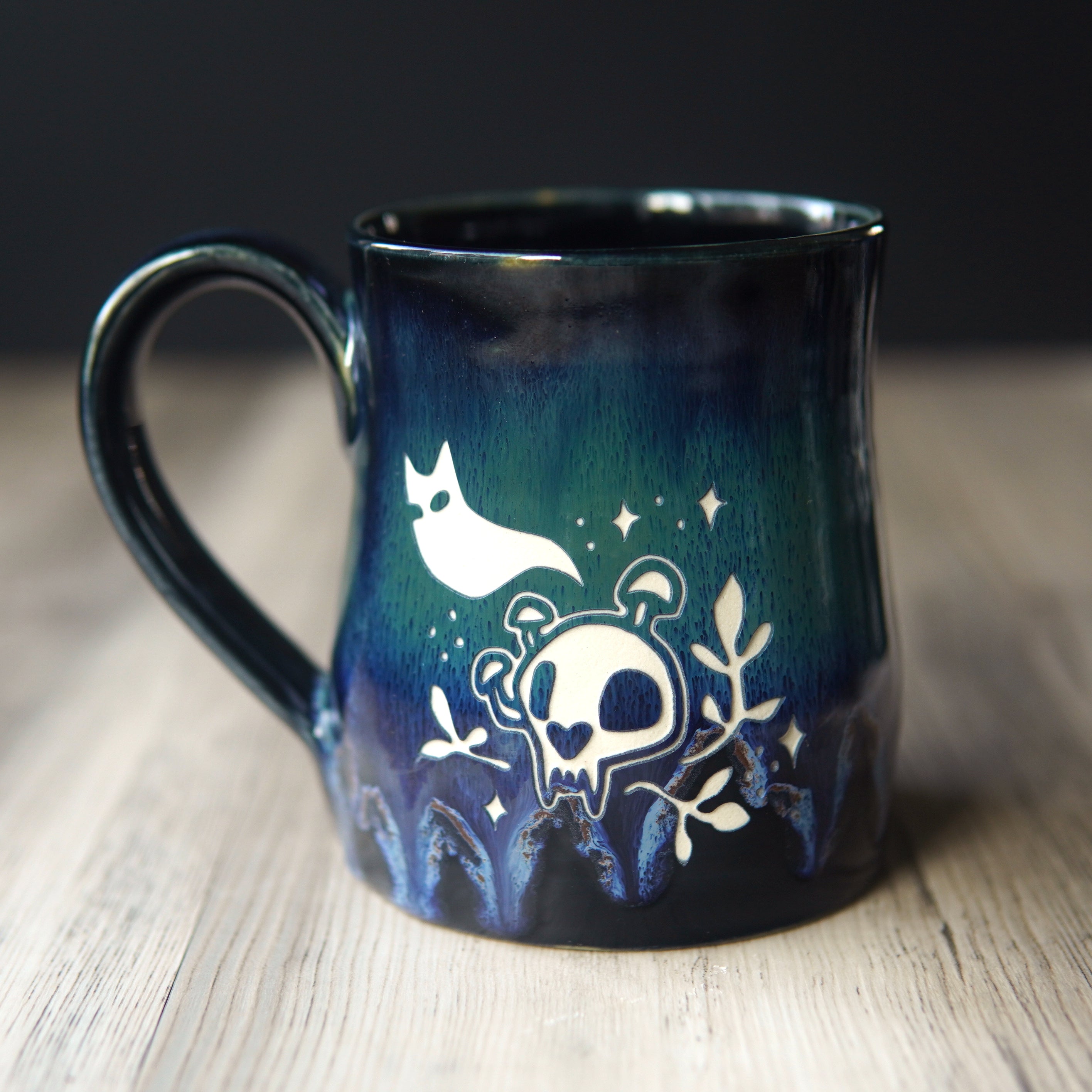 A dark blue and black glazed ceramic mug with a handle, decorated with a white skull, cat ghost, and stars design, resting on a light wood surface.