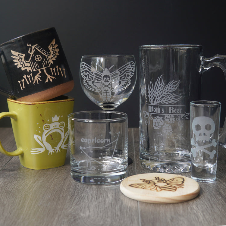 Collection of engraved mugs, glasses, and coasters on a wooden surface with a dark background.
