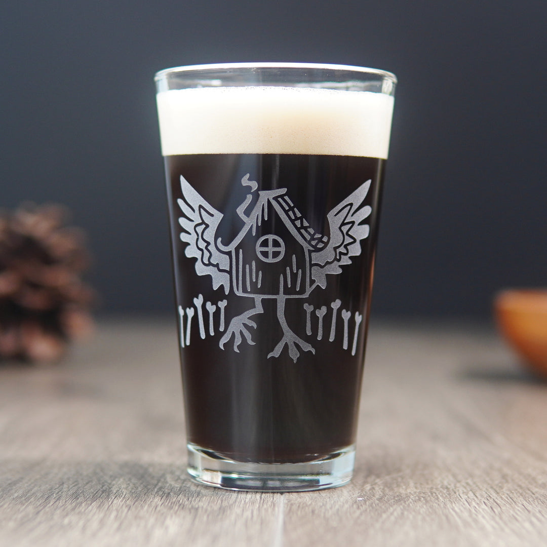 pint glass engraved with baba yaga's hut, with outspread chicken wings and legs, and a fence of bones. The glass is full of a dark beer.