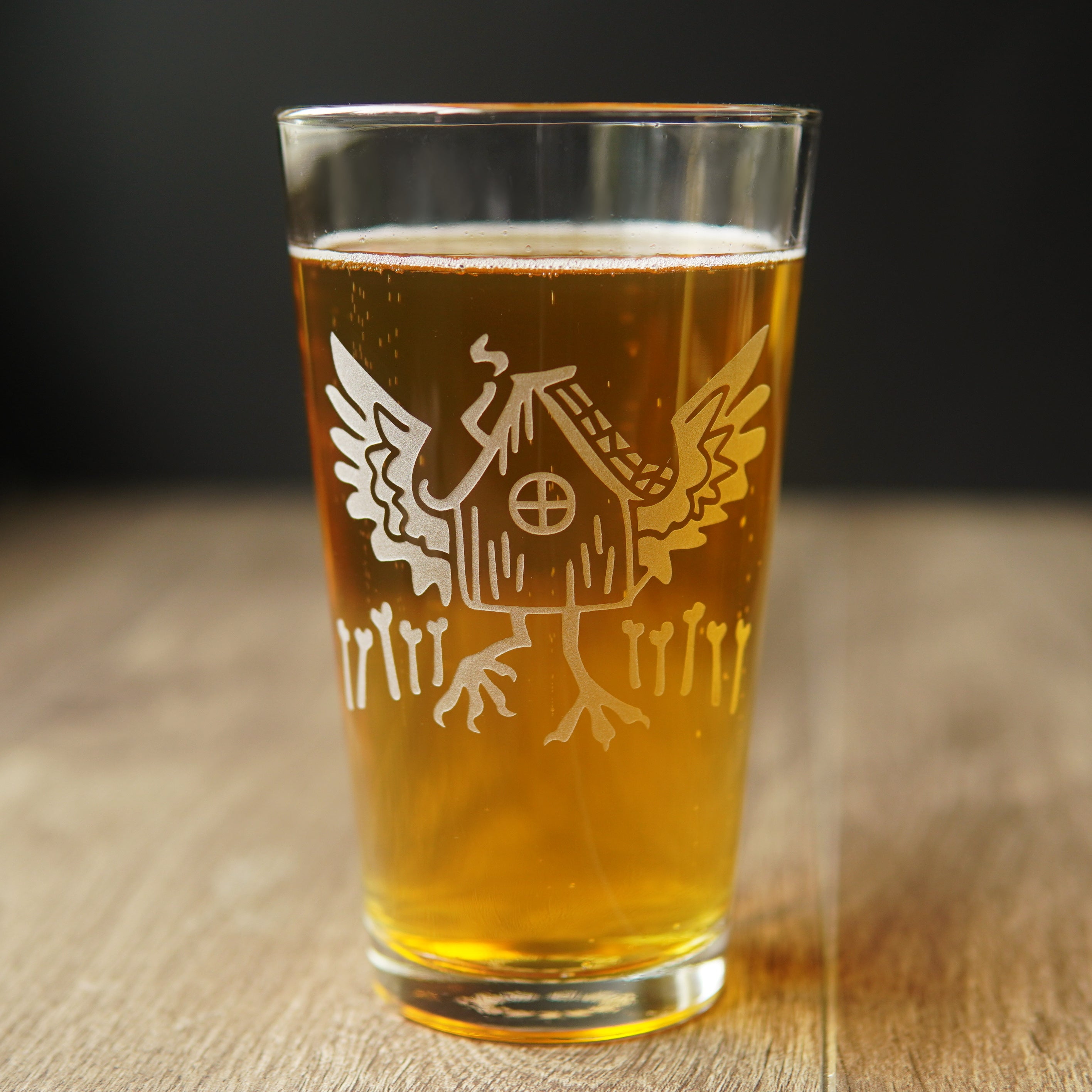 pint glass engraved with baba yaga's hut, with outspread chicken wings and legs, and a fence of bones. The glass is full of an amber liquid.