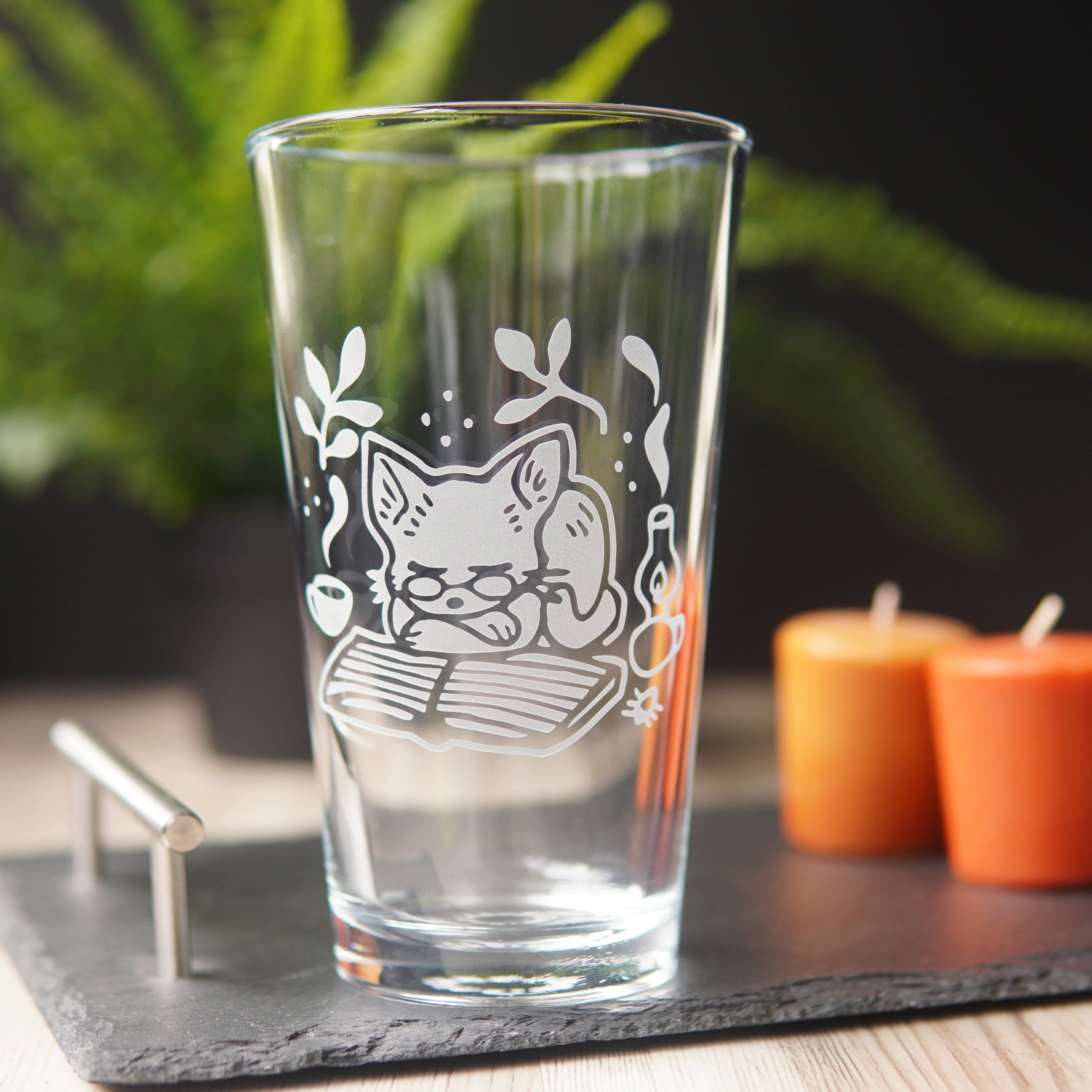 pint glass engraved with a cute cat. The cat is wearing glasses, and is curled up asleep on a book next to a lantern and steaming mug.