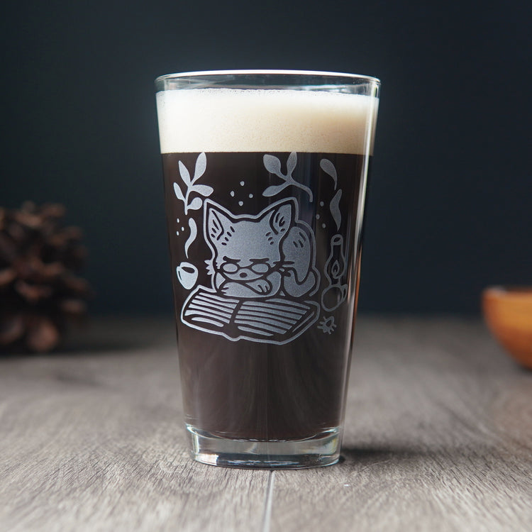 pint glass engraved with a cute cat. The cat is wearing glasses, and is curled up asleep on a book next to a lantern and steaming mug.