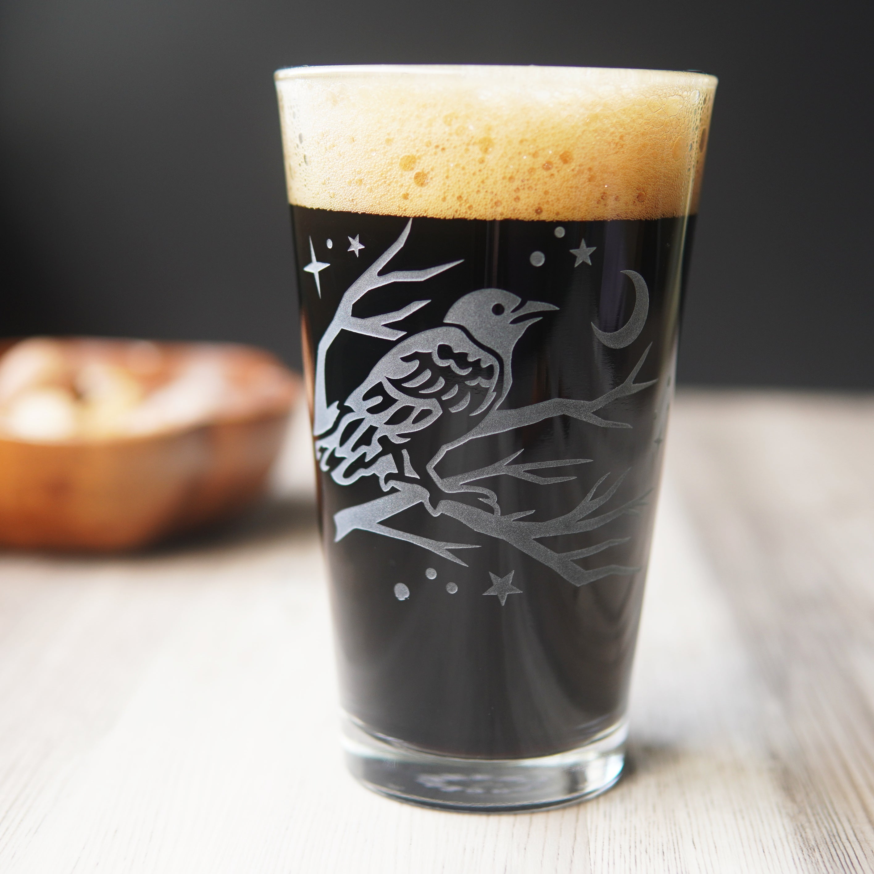 pint glass full of dark beer etched with a crow in a tree, stars, and a crescent moon