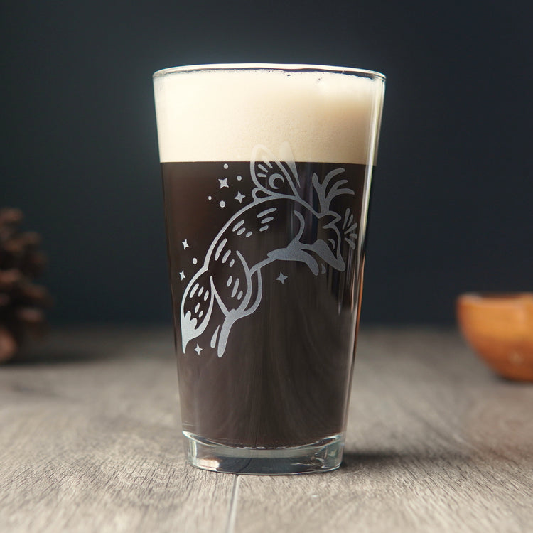 Pint glass etched with a leaping fox, full of dark beer. The fox has small butterfly wings and antlers, and is surrounded by sparkles.