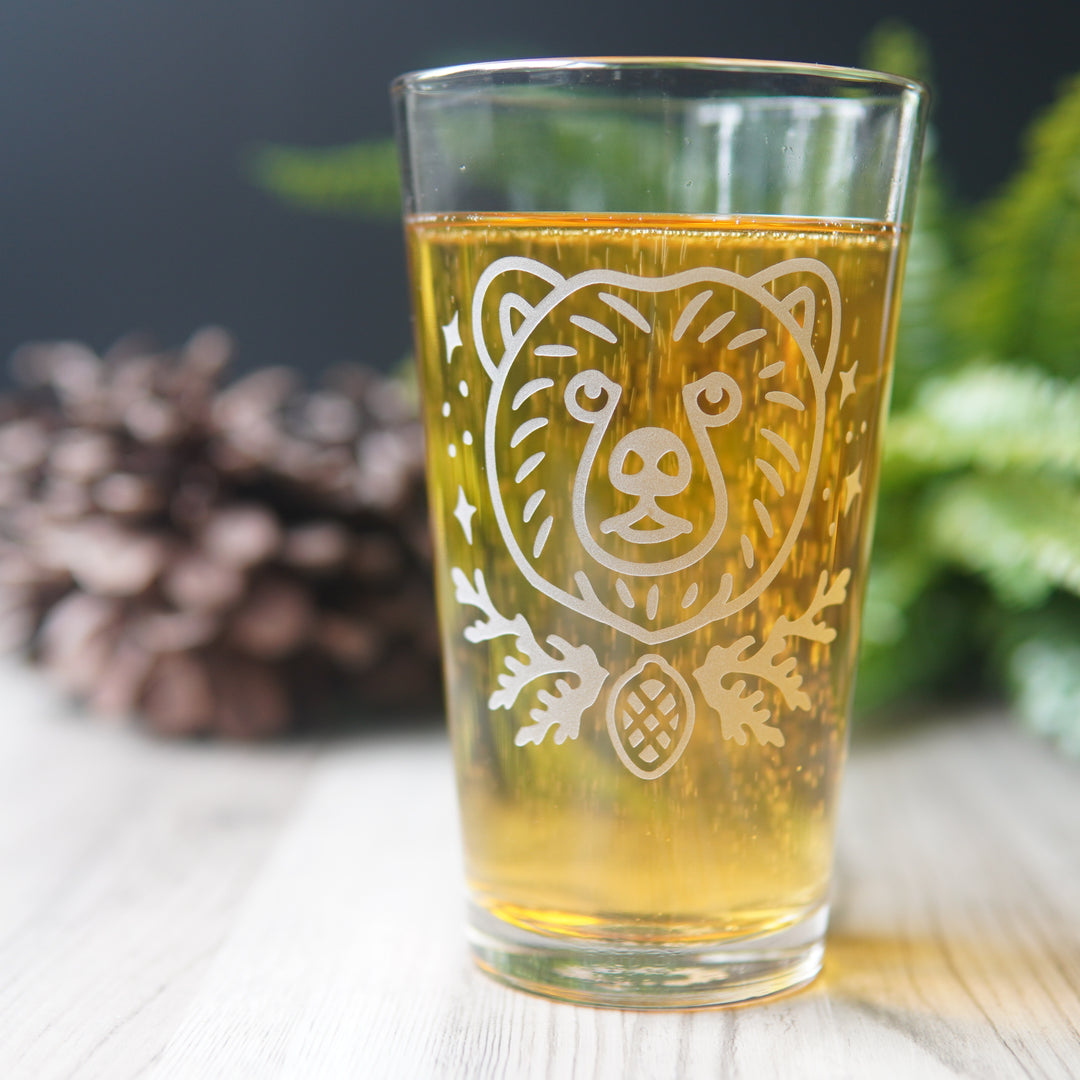 pint glass engraved with a bear's face, stars, cedar branches, and a pine cone