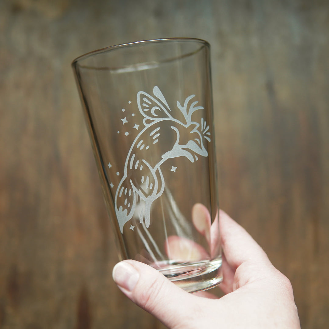 Pint glass etched with a leaping fox, held in a hand. The fox has small butterfly wings and antlers, and is surrounded by sparkles.