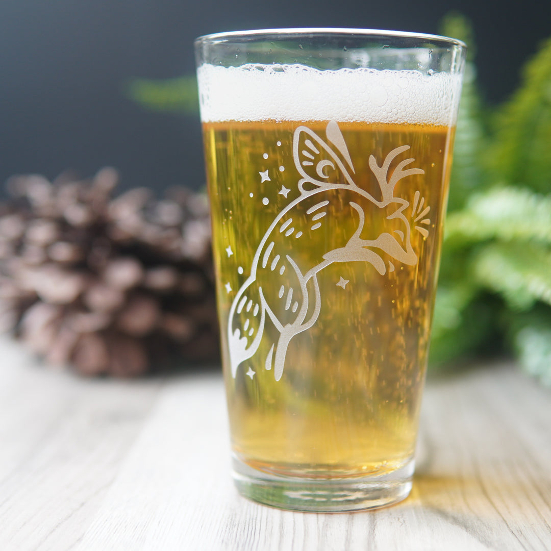 Pint glass etched with a leaping fox fairy. The fox has small butterfly wings and antlers, and is surrounded by sparkles.