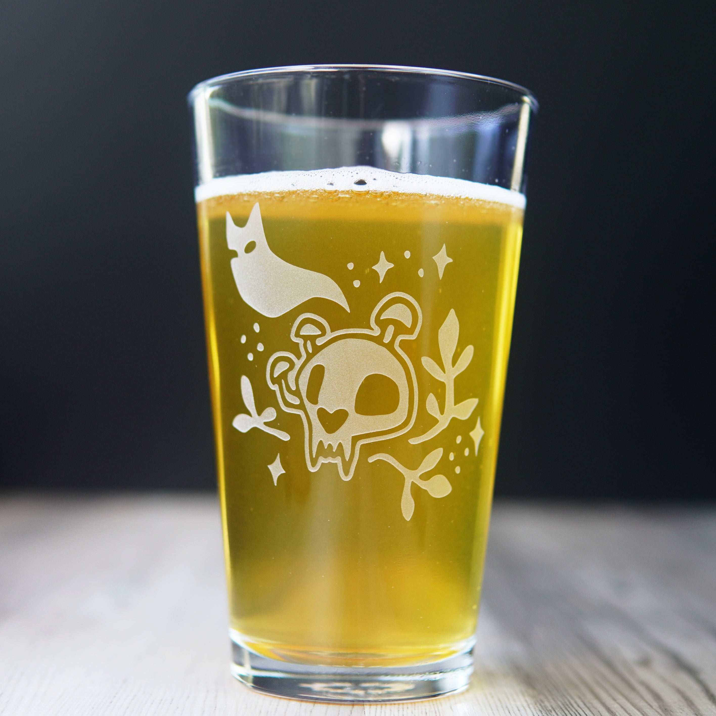 Pint glass full of beer engraved with a cartoonish cat skull with mushrooms growing on it. A cat ghost floats above, and sparkles and plants surround the skull.