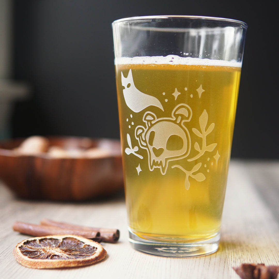 Pint glass engraved with a cat skull with mushrooms growing on it. A cat ghost floats above, and sparkles and plants surround the skull.