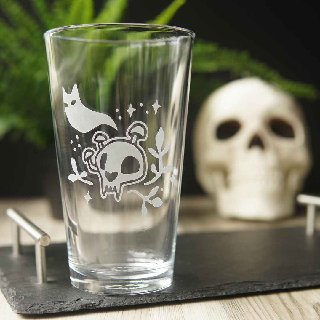 Pint glass engraved with a cat skull with mushrooms growing on it. A cat ghost floats above, and sparkles and plants surround the skull.