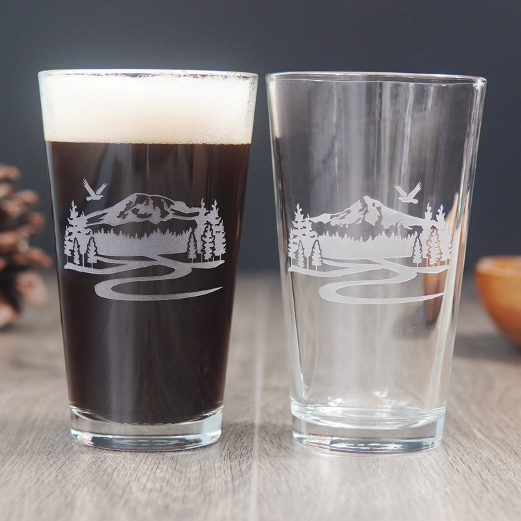 Pair of etched pint glasses. The one on the left is engraved with a scene of Mt Rainier, and the one of the right is Mt Hood. Both designs have trails below the mountains, fir trees on either side, and a bald eagle flying overhead.