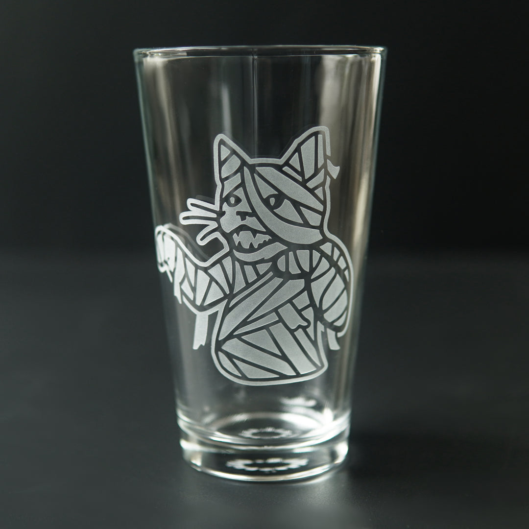 pint glass etched with the upper body of a mummy cat. The cat's mouth is open, arms slightly outstretched, and wrappings are coming loose.