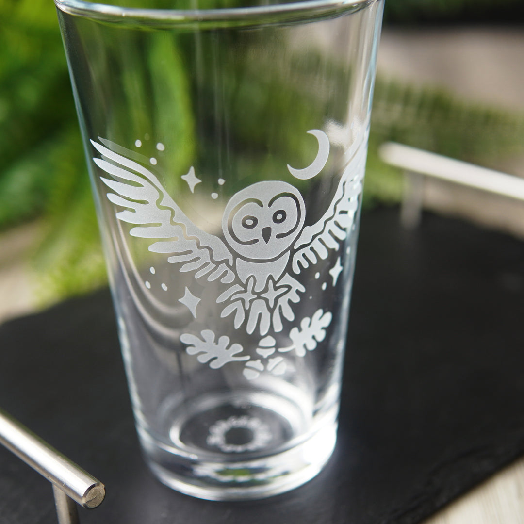 etched pint glass with a flying great grey owl surrounded by stars. A crescent moon hangs above the owl, and oak leaves and acorns float below its open claws