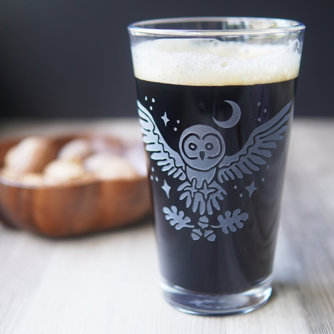 etched pint glass with a flying great grey owl surrounded by stars. A crescent moon hangs above the owl, and oak leaves and acorns float below its open claws