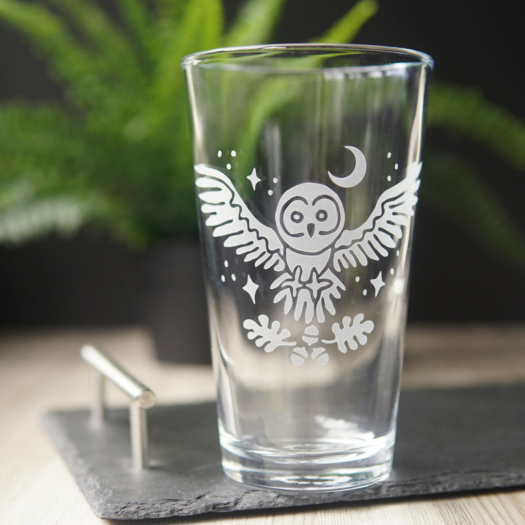 etched pint glass with a flying great grey owl surrounded by stars. A crescent moon hangs above the owl, and oak leaves and acorns float below its open claws