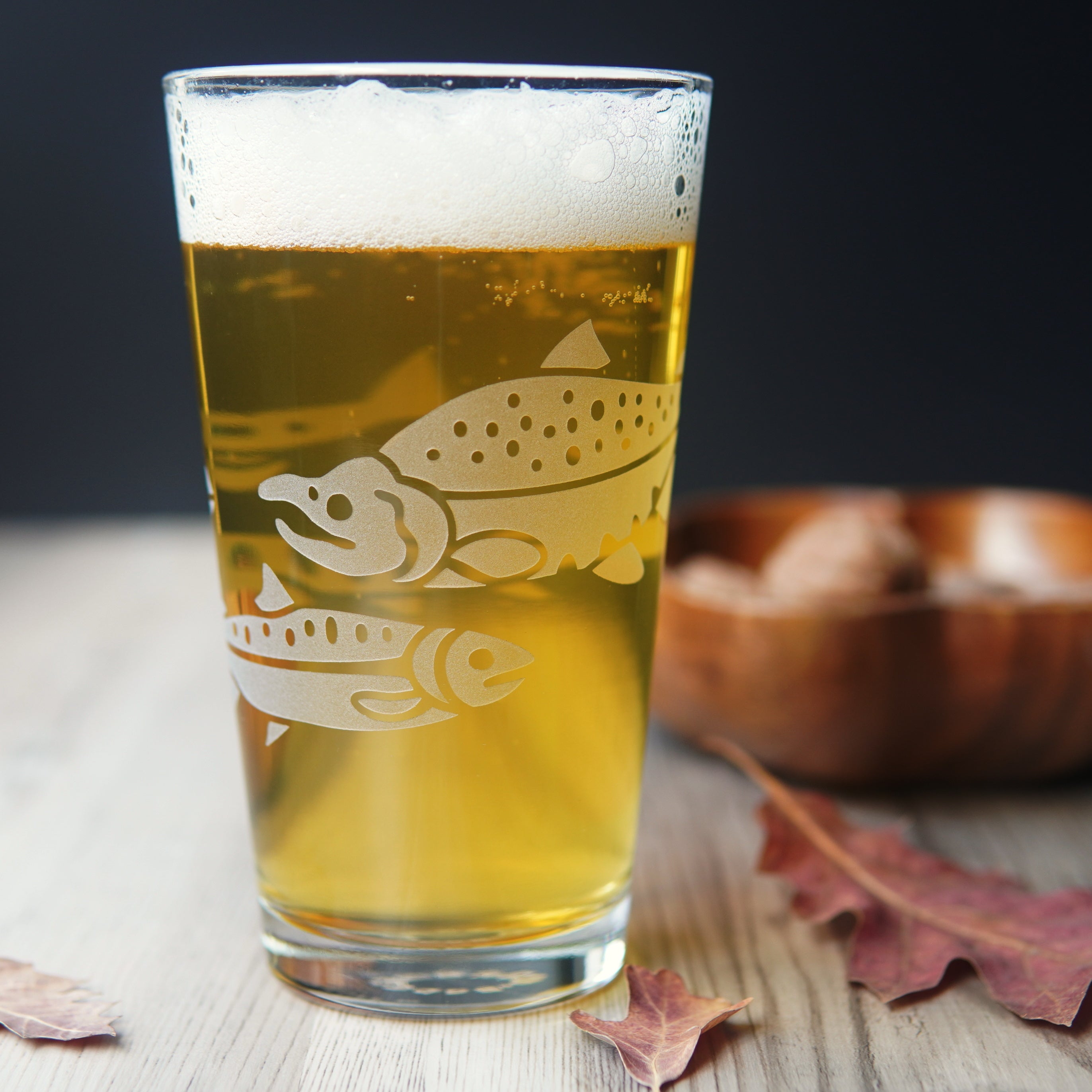 Salmon pint glass full of yellow beer, sitting next to red oak leaves and a wooden bowl of nuts