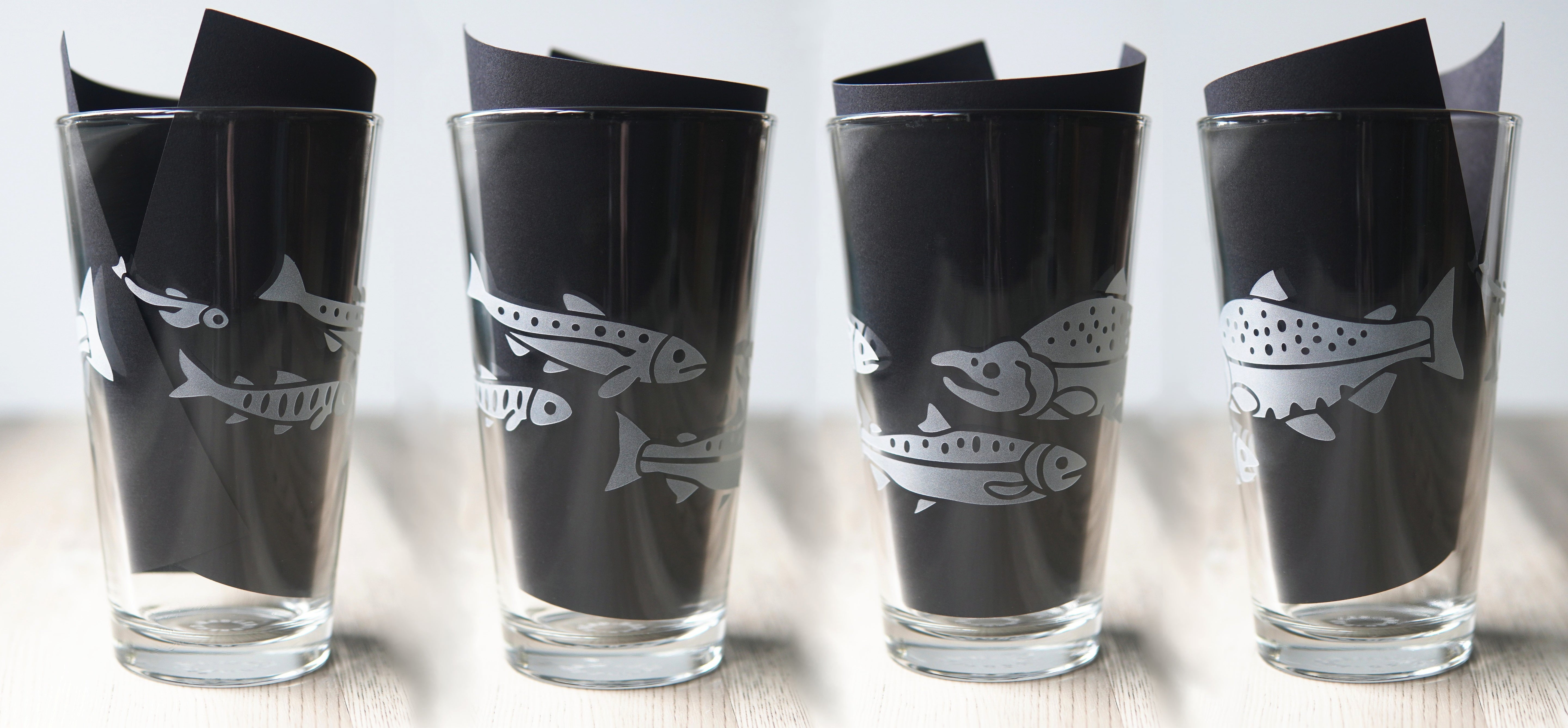 4 pint glasses etched with salmon. The glasses show different angles of the same design, with fish at all stages of their life cycle