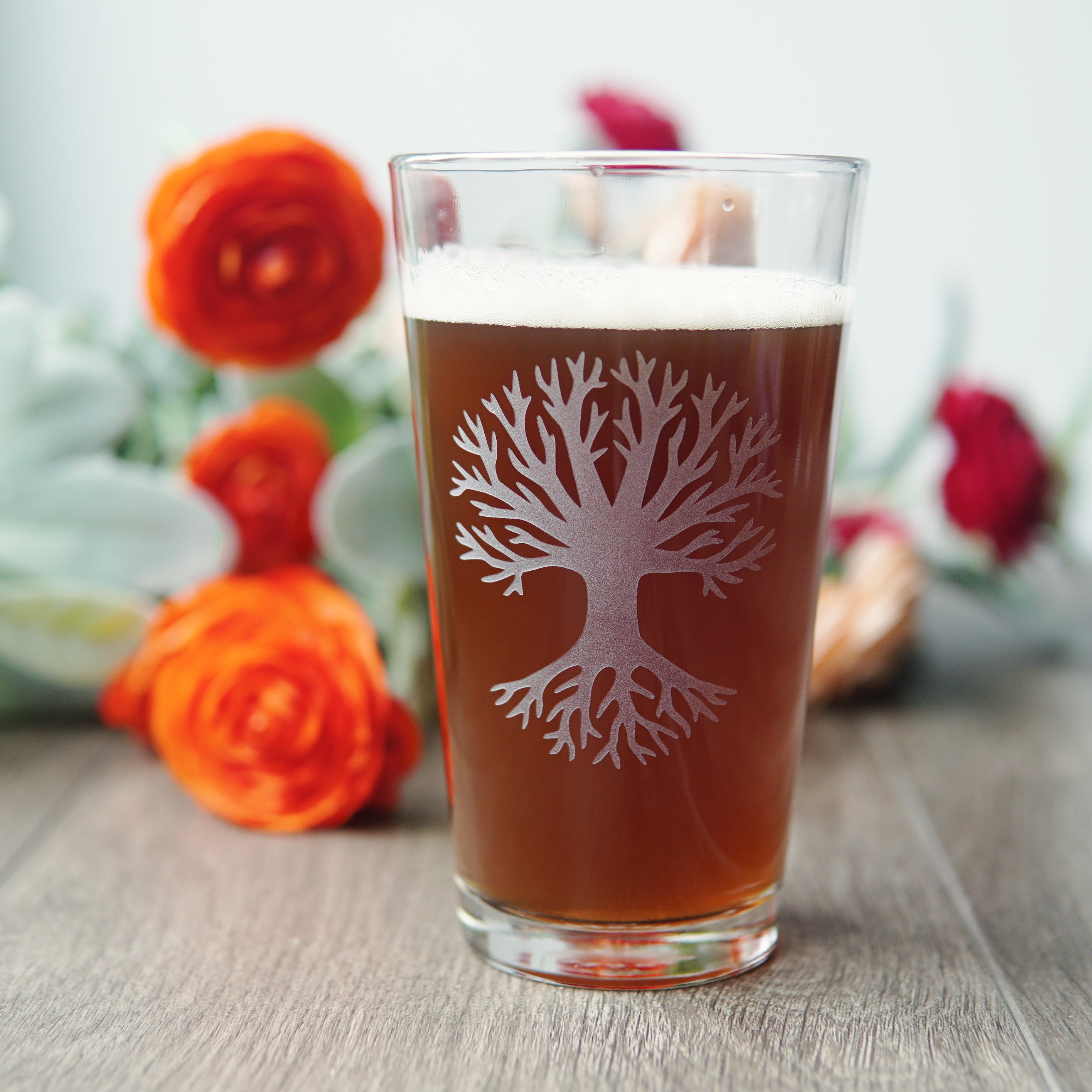 pint glass etched with a large bare tree and wide roots, sitting in front of bright ranunculus flowers