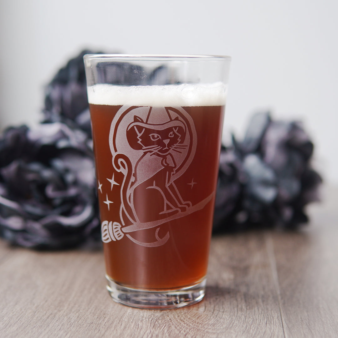 pint glass engraved with a witch cat on a flying broom in front of the full moon and stars. The glass is full of red beer or kombucha, sitting in front of black flowers