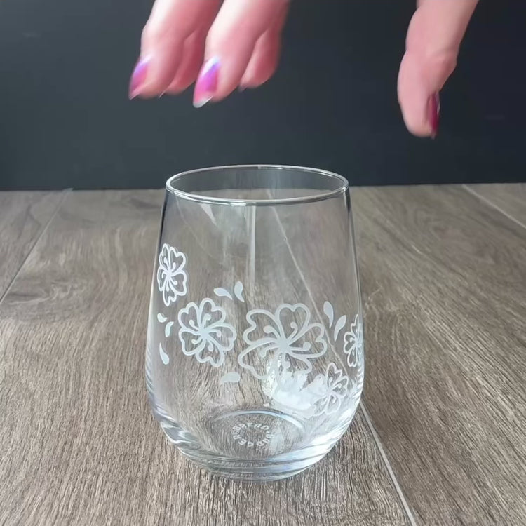 Sped up video of a hand manipulating an etched stemless wine glass so it can be viewed from all angles. Then sped up clips of a woman masking a glass, then standing in front of a sandblasting cabinet putting on a respirator, then a view of the inside of the cabinet showing a glass being sandblasted.