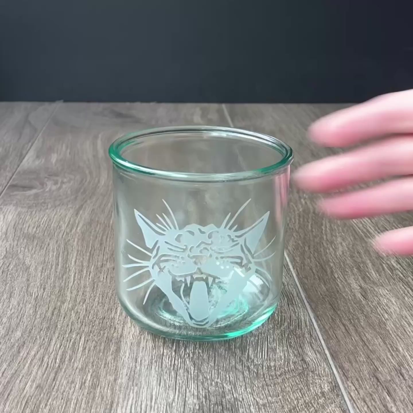 sped up video of a hand manipulating an etched short glass tumbler, then a tall glass tumbler, so they can be viewed from all angles. Then sped up clips of a woman masking a glass, then standing in front of a sandblasting cabinet putting on a respirator, then a view of the inside of the cabinet showing a glass being sandblasted.
