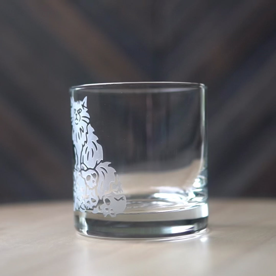 Sped up video of a hand manipulating a lowball cocktail glass so it can be viewed from all angles. Then sped up clips of a woman masking a glass, then standing in front of a sandblasting cabinet putting on a respirator, then a view of the inside of the cabinet showing a glass being sandblasted.
