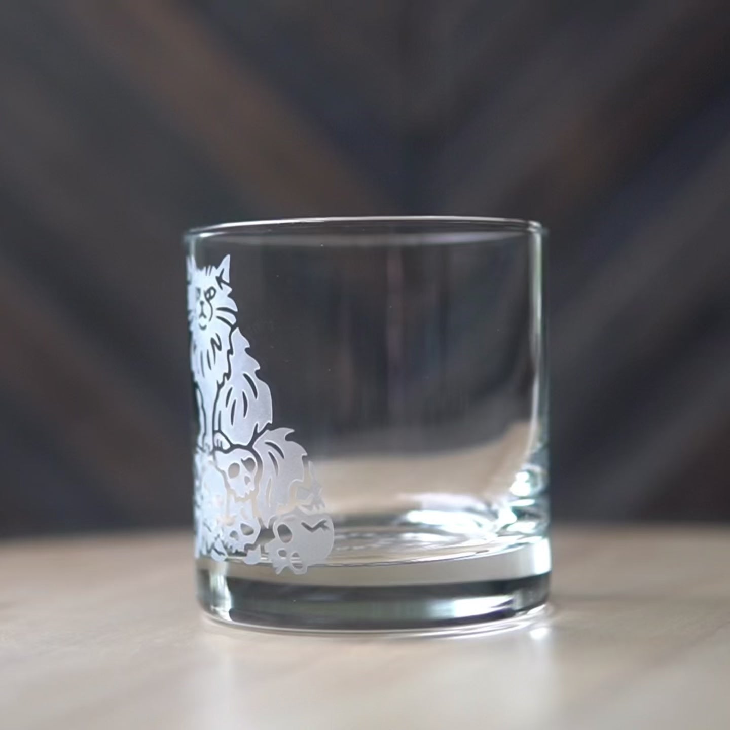 Sped up video of a hand manipulating a lowball cocktail glass so it can be viewed from all angles. Then sped up clips of a woman masking a glass, then standing in front of a sandblasting cabinet putting on a respirator, then a view of the inside of the cabinet showing a glass being sandblasted.
