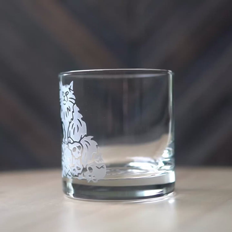 Sped up video of a hand manipulating a lowball cocktail glass so it can be viewed from all angles. Then sped up clips of a woman masking a glass, then standing in front of a sandblasting cabinet putting on a respirator, then a view of the inside of the cabinet showing a glass being sandblasted.