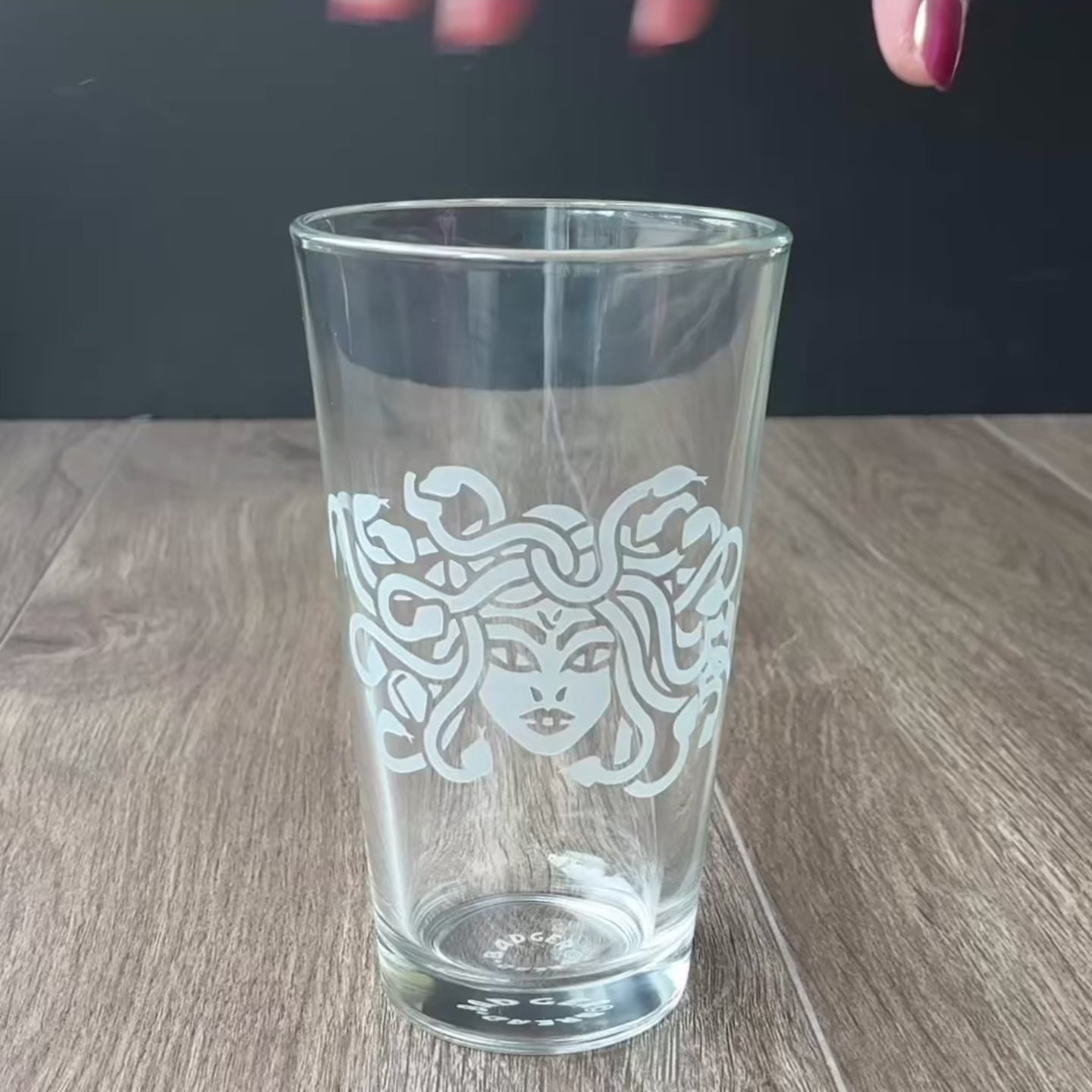 Sped up video of a hand manipulating an etched pint glass so it can be viewed from all angles. Then sped up clips of a woman masking a glass, then standing in front of a sandblasting cabinet putting on a respirator, then a view of the inside of the cabinet showing a glass being sandblasted.