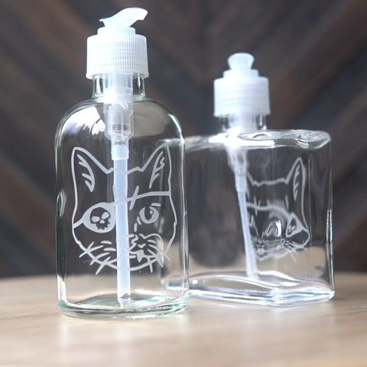 Video of two etched glass soap dispensers, rotating on a lazy susan so they can be viewed from all angles. Then sped up clips of a woman masking a glass, then standing in front of a sandblasting cabinet putting on a respirator, then a view of the inside of the cabinet showing a glass being sandblasted.