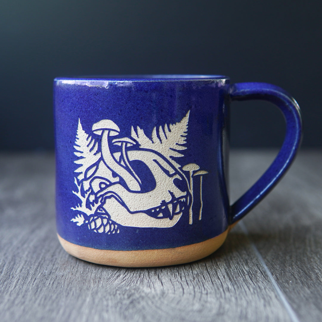 skull mug with cobalt blue glaze. the skull is from a cat, and has mushrooms growing out of the eye socket, with ferns behind it.