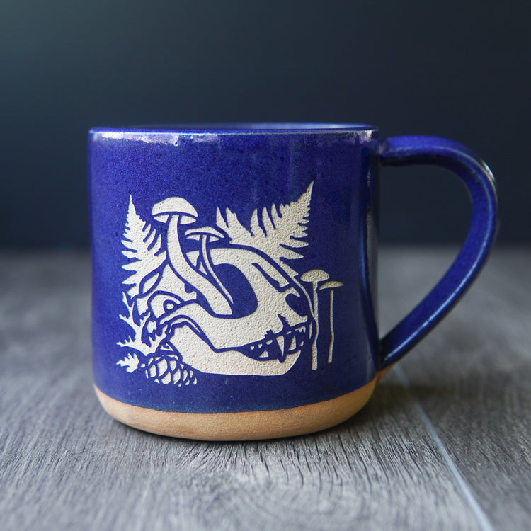skull mug with cobalt blue glaze. the skull is from a cat, and has mushrooms growing out of the eye socket, with ferns behind it.