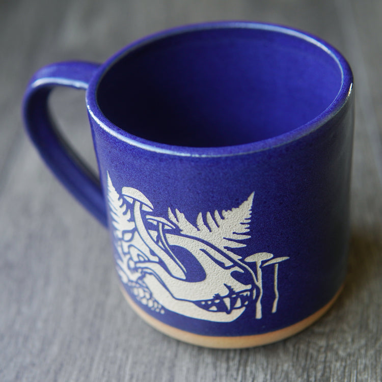 skull mug with cobalt blue glaze. the skull is from a cat, and has mushrooms growing out of the eye socket, with ferns behind it.