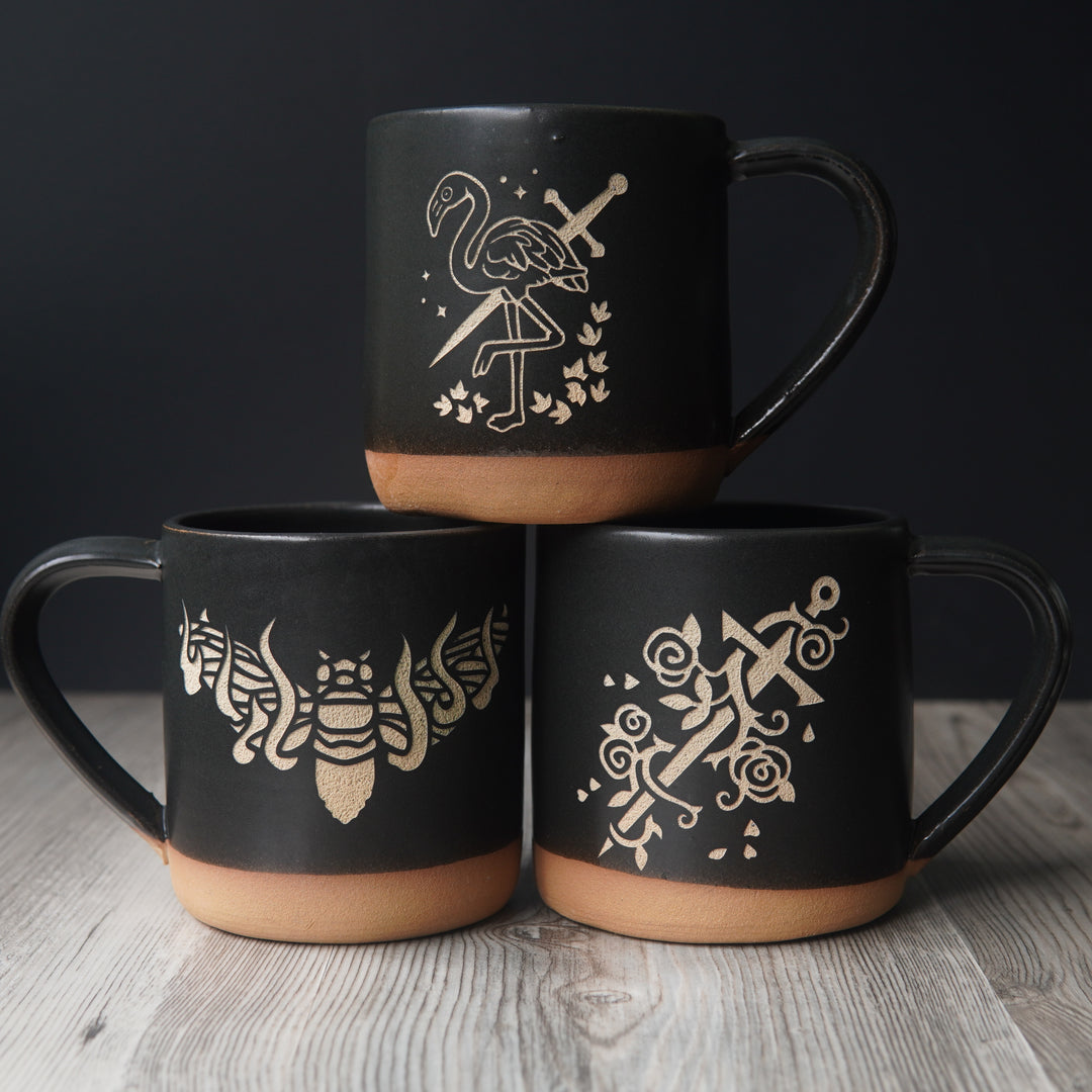 black mugs: flamingo with a sword, flaming cicada, and sword with rose vines