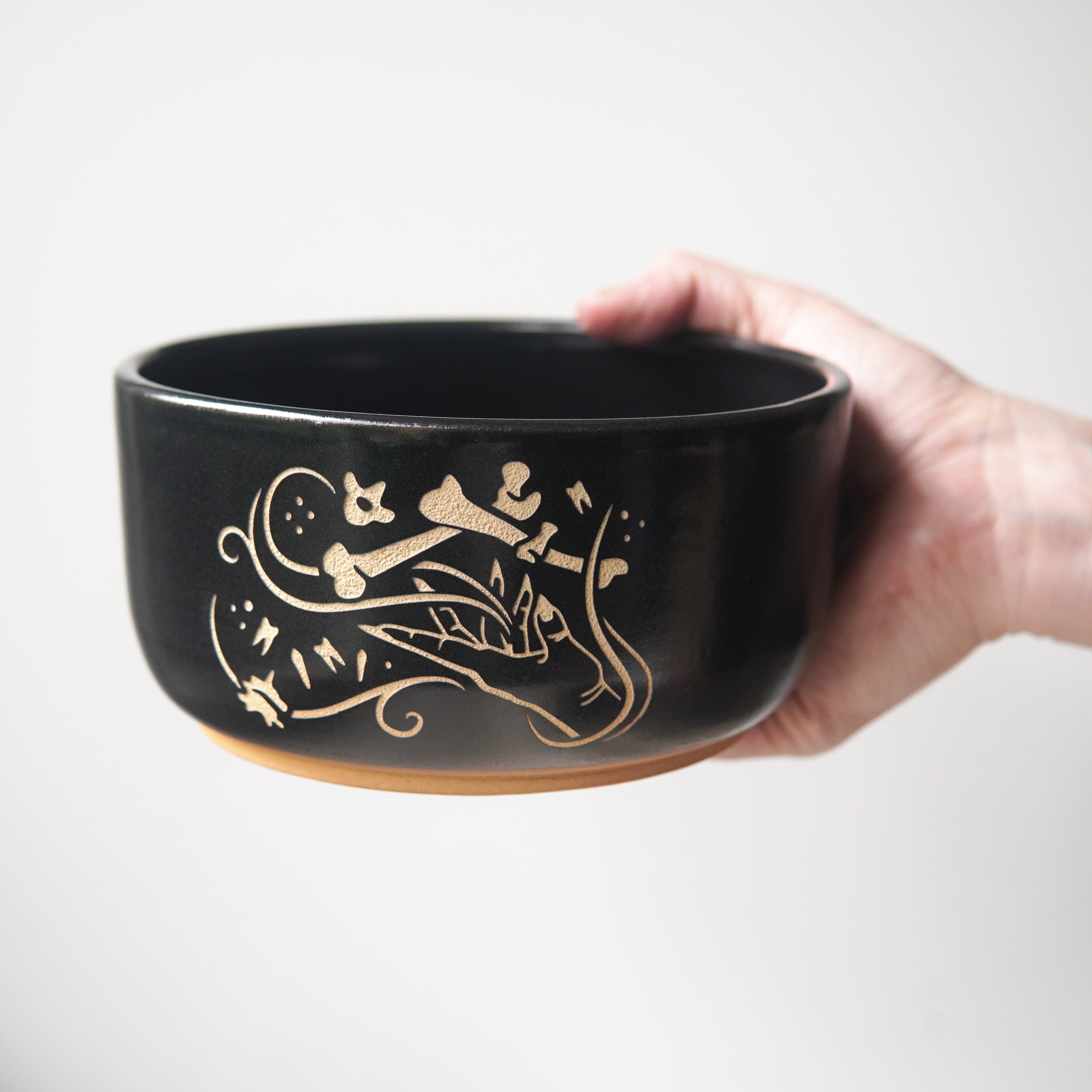 Ramen sized black pottery bowl held by a hand. The bowl has an engraved graphic of a hand levitating human bones and teeth with swirls of magic