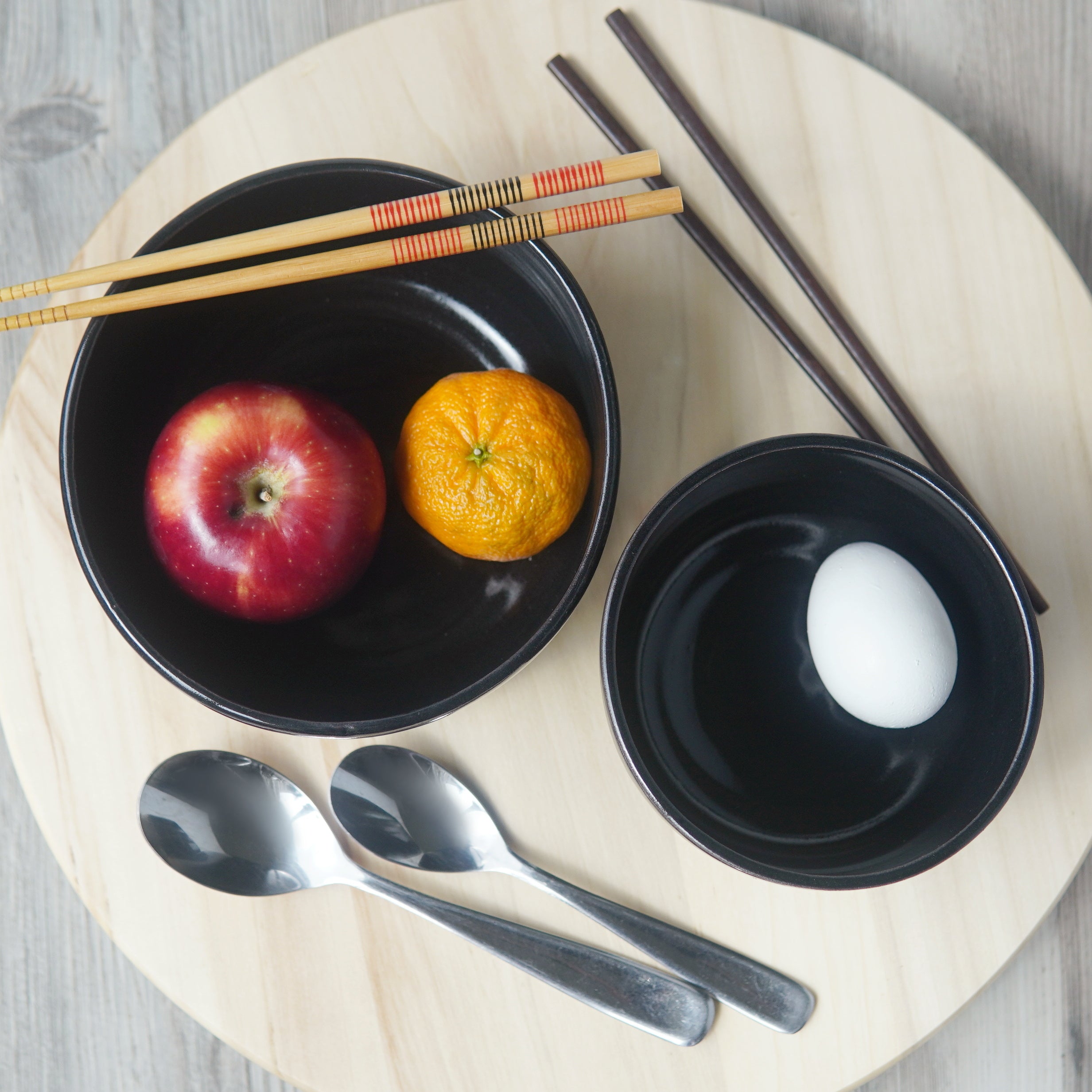 Two black pottery bowls, viewed from above. There are chopsticks and spoons next to the bowls. An apple, a small orange, and an egg are inside the bowls.