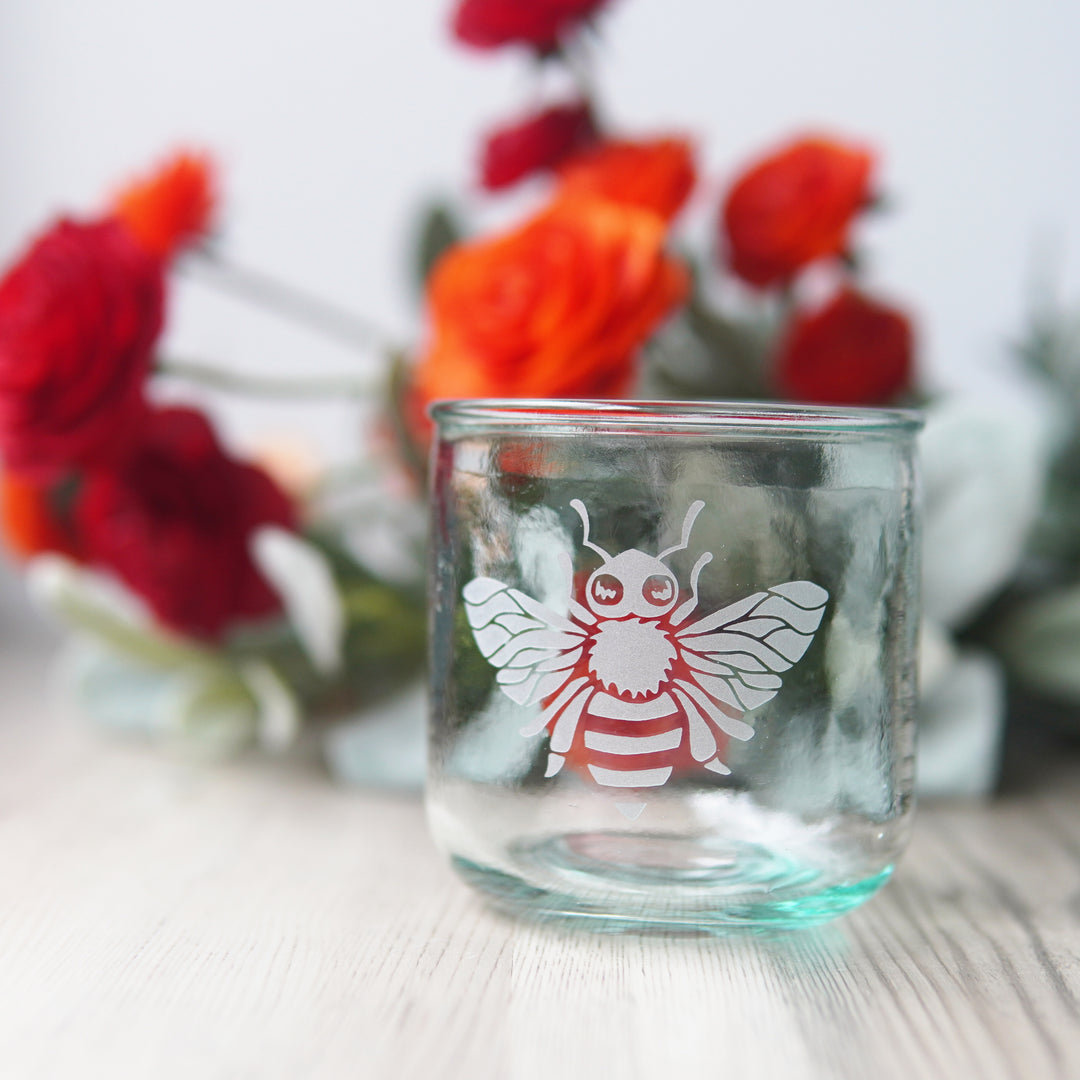 short recycled glass cup engraved with a honey bee, with flowers in the background