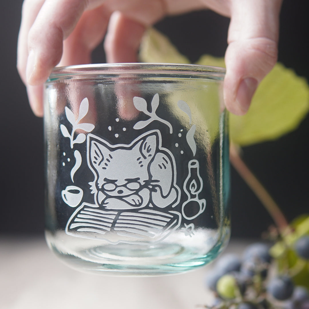 short recycled glass cup engraved with a cat wearing glasses, sleeping on a book. There is a steaming mug and lit lantern next to the cat.