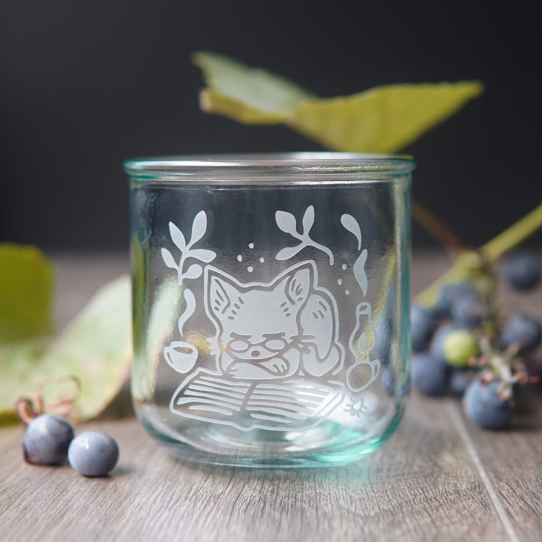 short recycled glass cup engraved with a cat wearing glasses, sleeping on a book. There are small grapes and vines behind the glass.