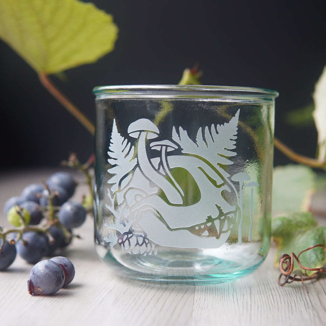 decaying cat skull with mushrooms and ferns etched onto a short glass. Grapes and vines are in the background.