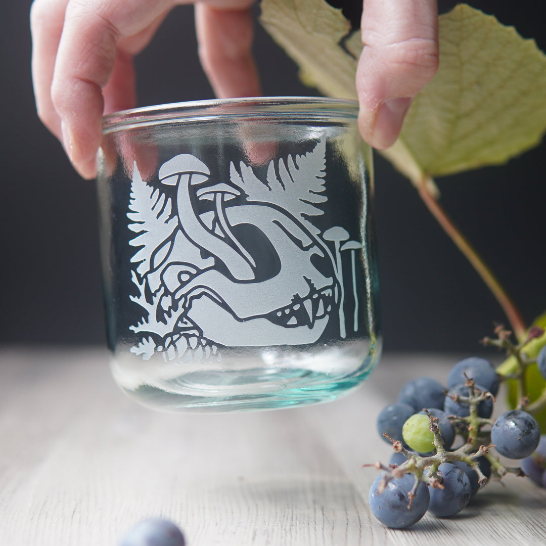 decaying cat skull with mushrooms and ferns etched onto a short glass held in a hand