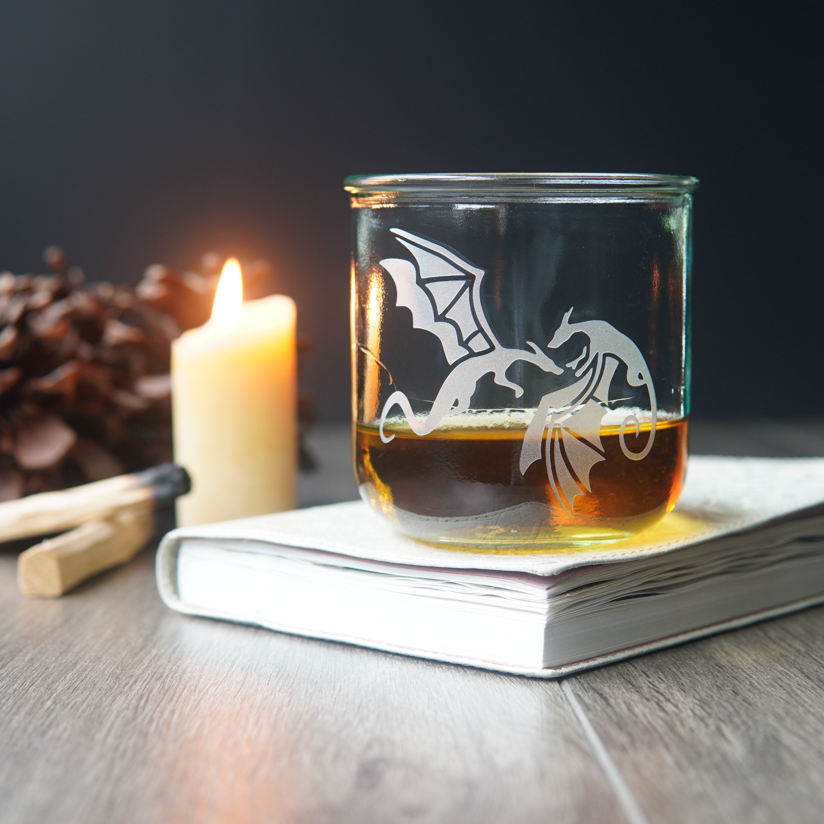 winged dragons etched on a short glass of whiskey, sitting on a book next to a lit candle