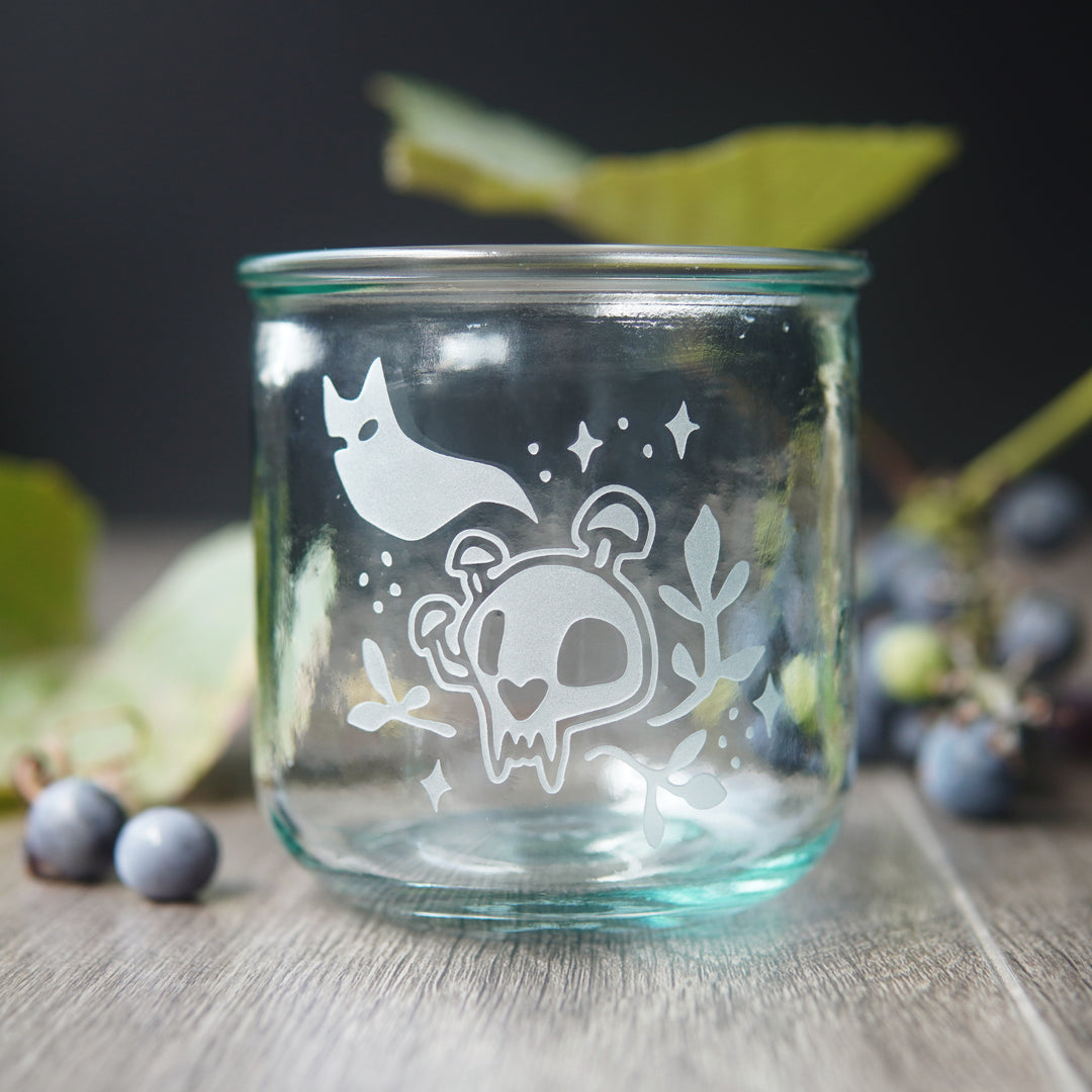 short glass engraved with a simplified cat skull with mushrooms growing on it, a cat ghost, and sparkles and plants surrounding it