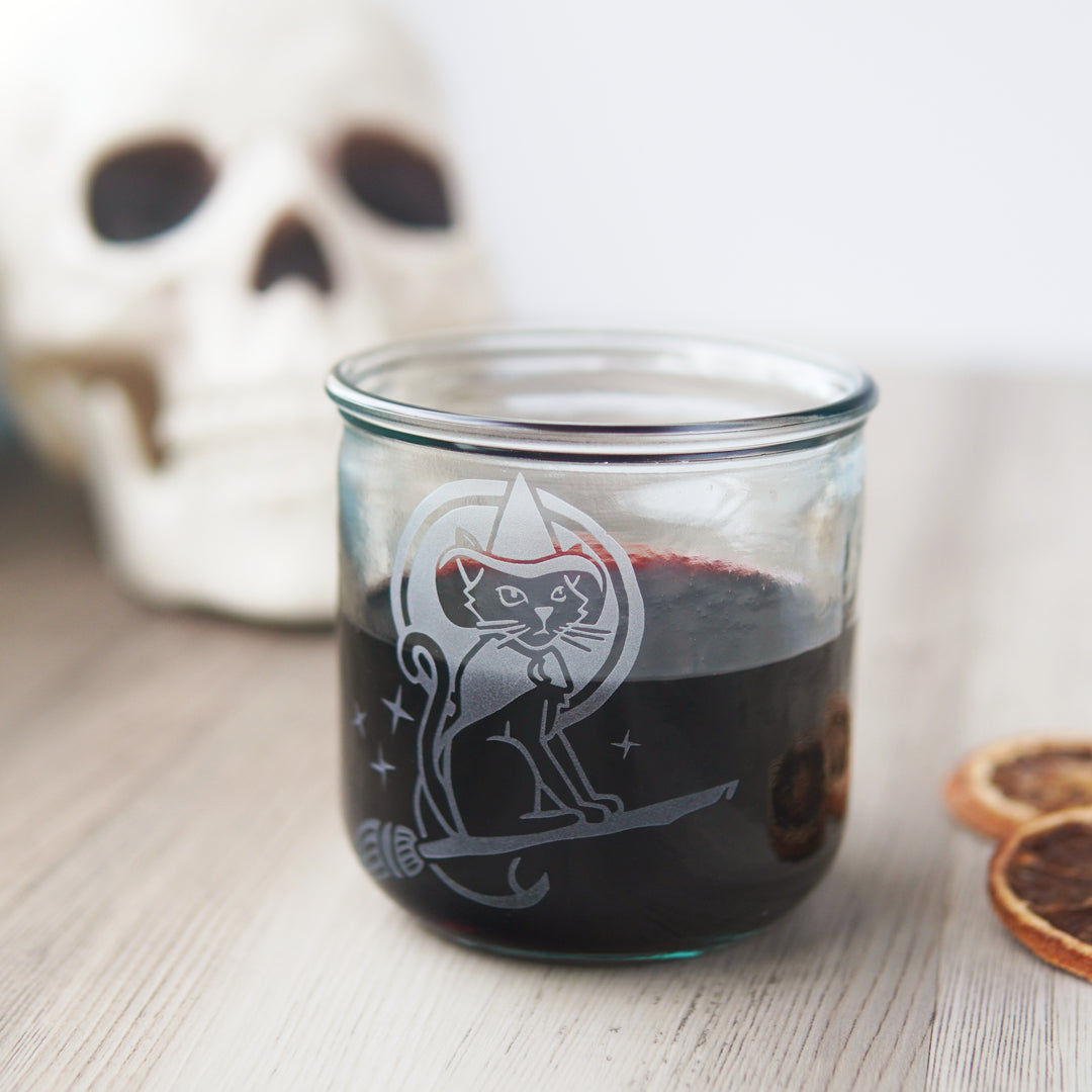 witch cat engraved onto a short glass with red wine, next to a plastic skull