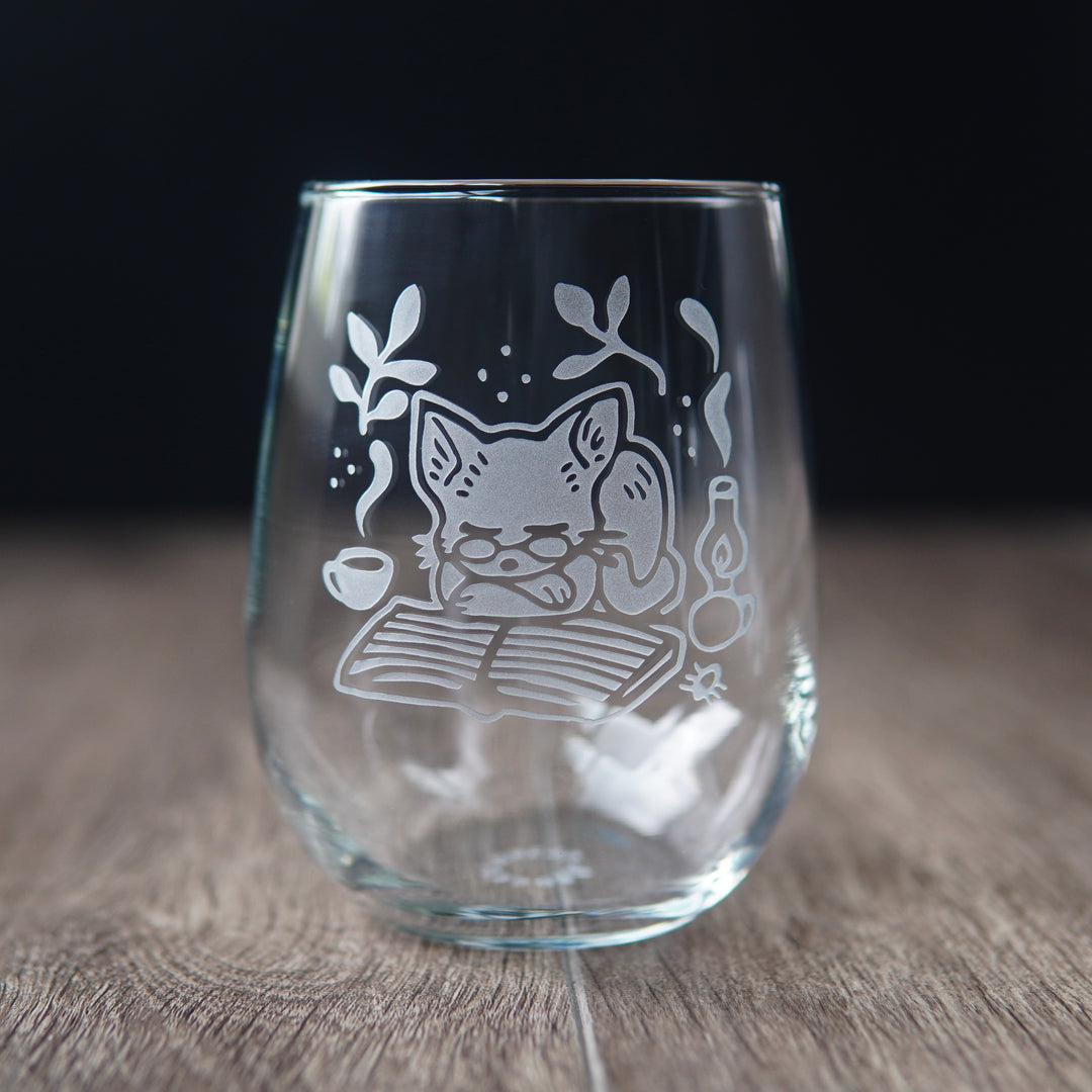 stemless wine glass etched with a sleeping cat on top of a book, wearing glasses, next to a steaming mug and oil lamp.