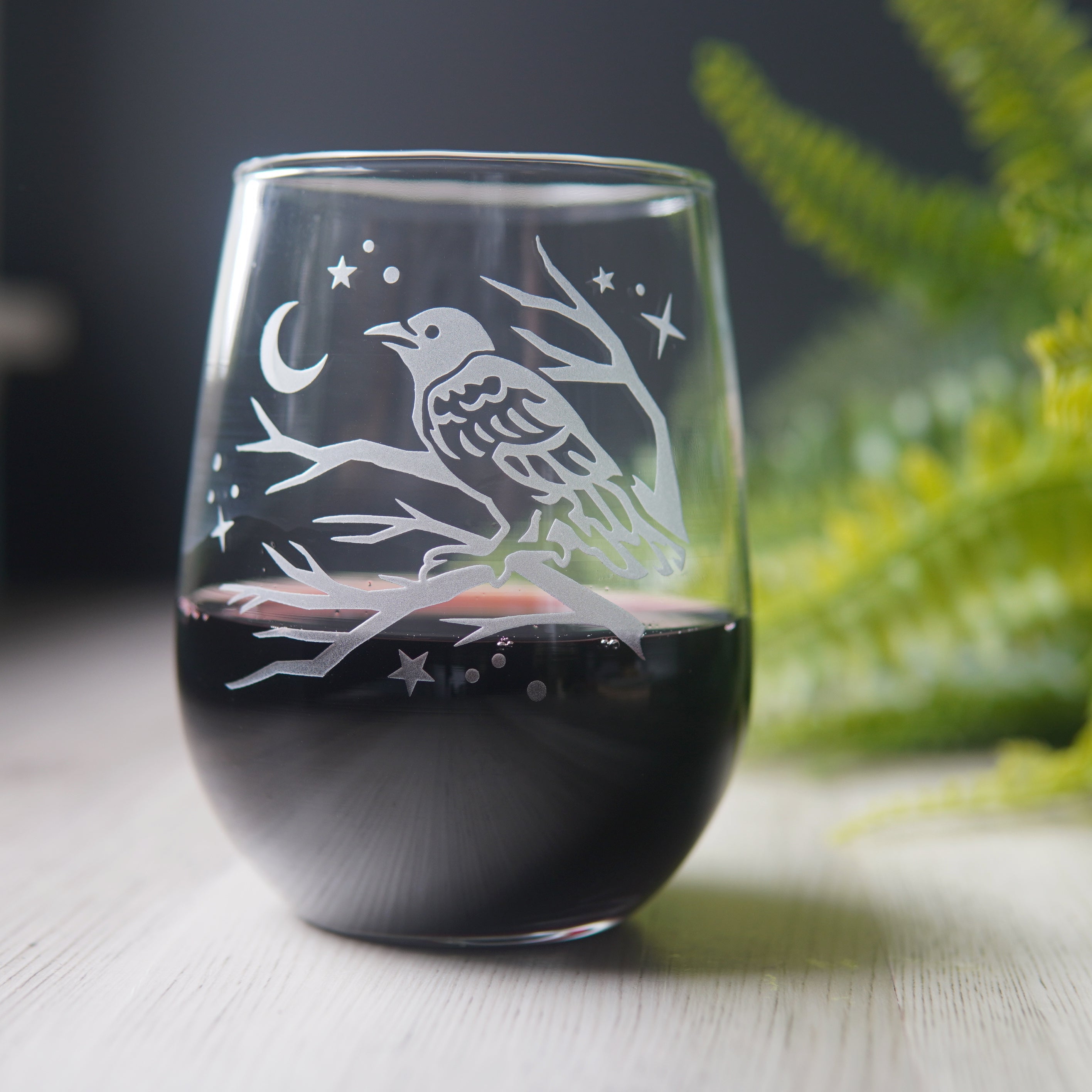 stemless wine glass etched with a crow in a tree, facing a crescent moon and stars. The glass has red wine in it and a fern in the background.