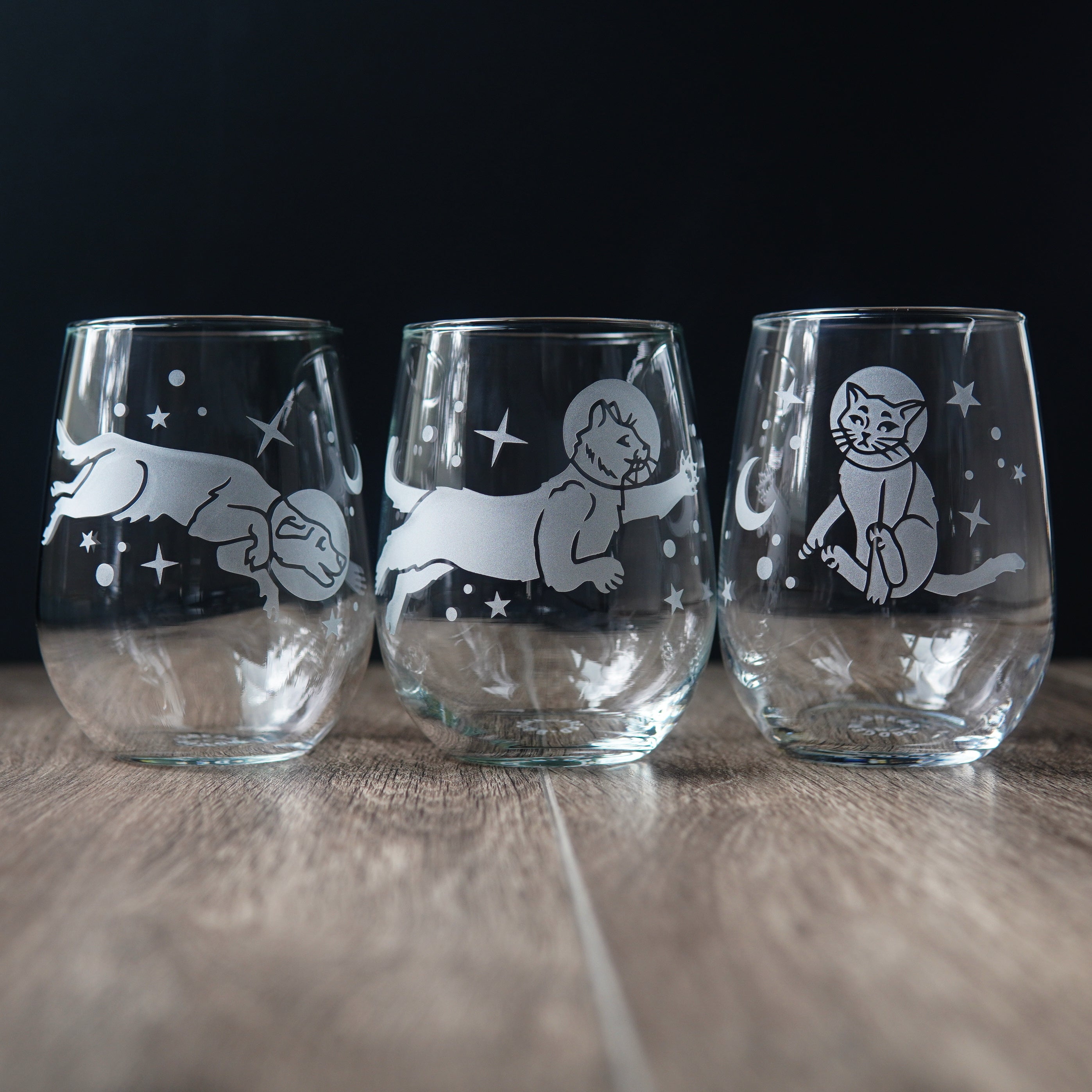 Three stemless wine glasses engraved with a dog and two different cats floating through the stars.