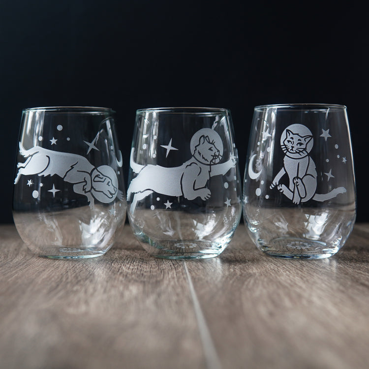 Three stemless wine glasses engraved with a dog and two different cats floating through the stars.