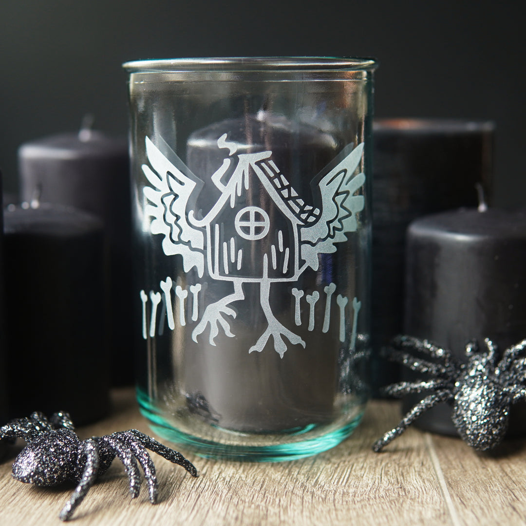 Baba Yaga's Hut etched onto a tall recycled glass cup/candle holder. Black candles and glittery plastic spiders surround it.